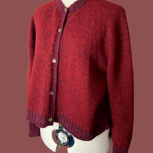 Puede incluir: Un cárdigan de punto rojo con cierre de botones y un ribete burdeos contrastante alrededor del cuello, los puños y el dobladillo.