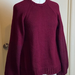 May include: A maroon knitted sweater with long sleeves and a round neckline. The sweater has a ribbed texture and a slightly loose fit.