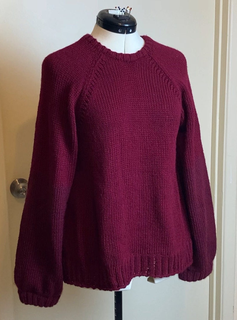 May include: A maroon knitted sweater with long sleeves and a round neckline. The sweater has a ribbed texture and a slightly loose fit.