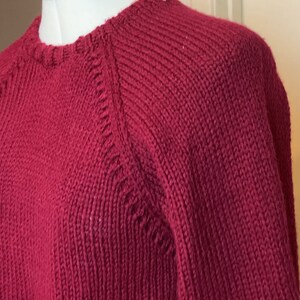 May include: A maroon knitted sweater with a round neckline and long sleeves. The sweater has a ribbed texture and a slightly loose fit.