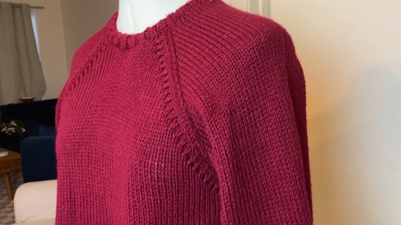 May include: A maroon knitted sweater with a round neckline and long sleeves. The sweater has a ribbed texture and a slightly loose fit.