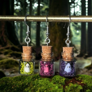 以下が含まれることがあります： コルクの蓋と銀色のフックが付いたガラス瓶のイヤリングが3つ。それぞれの瓶には小さな幽霊の置物と異なる色の苔が入っています。幽霊は白、ピンク、紫です。