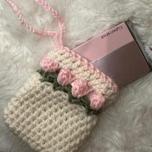 May include: A white crocheted camera case with pink and green trim. The case has a pink strap and is holding a pink and silver camera with the text "Cyber-shot" on it.