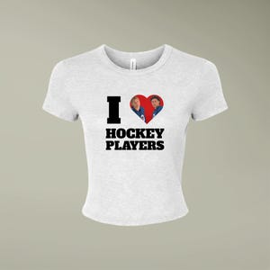 Puede incluir: Una camiseta gris claro de manga corta con el texto negro I HOCKEY PLAYERS, donde un corazón rojo sustituye a la letra O. Dentro del corazón aparecen dos personas con camisetas de hockey azules, una al lado de la otra.