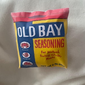 May include: A yellow, blue, and red bag of Old Bay Seasoning. The bag features the words "OLD BAY SEASONING" in white and red, with illustrations of a crab, shrimp, and other seafood. The text "For Seafood, Poultry, Salads, Meats" is also visible.
