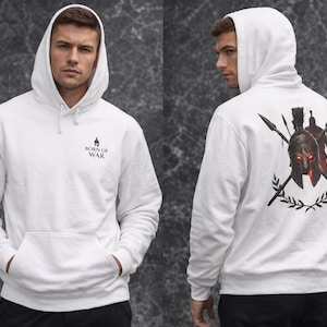 Puede incluir: Sudadera con capucha blanca con bolsillo delantero y capucha. La parte delantera presenta el texto "BORN OF WAR" y un pequeño gráfico. La parte trasera muestra un gráfico de un casco espartano, lanzas y hojas de laurel.