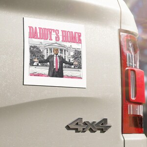 May include: A white square sticker with a black and white image of a man in a suit standing in front of the White House. The text "DADDY'S HOME" is in pink letters above the image.