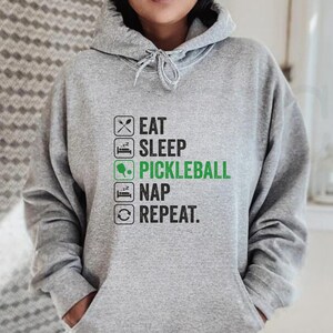 May include: A light gray hoodie with a drawstring hood and a front pocket. The text "EAT SLEEP PICKLEBALL NAP REPEAT" is printed in black and green. The person is wearing denim shorts.