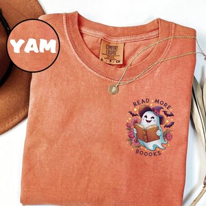 Op de afbeelding: Oranje Comfort Colors T-shirt met een afbeelding van een spook dat een boek leest, een heksenhoed draagt en omringd is door vleermuizen en bloemen. De tekst "Read More Boooks" staat boven en onder het spook.