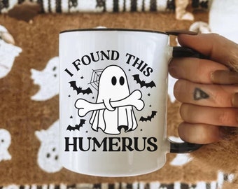 Op de afbeelding: Witte keramische mok met een zwarte rand en handvat. De mok heeft een Halloween-thema ontwerp met een spook dat een bot vasthoudt, vleermuizen en de tekst "I FOUND THIS HUMERUS".