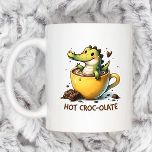 Pode incluir: Caneca de cerâmica branca com um jacaré de desenho animado numa chávena amarela de chocolate quente. O jacaré é verde e sorri. A caneca tem a inscrição "HOT CROC-OLATE". Há pedaços de chocolate ao lado da chávena.