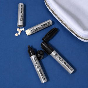Peut inclure: Deux marqueurs argentés Sharpie Magnum Size avec capuchons noirs, dont un sans capuchon, et deux corps de marqueur argentés supplémentaires. Un corps est ouvert avec de petites pilules beiges rondes éparpillées. Une pochette blanche est visible.