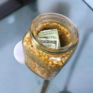 Peut inclure: Un bocal en verre rempli de grains de maïs jaunes, avec quelques billets de banque à l'intérieur.