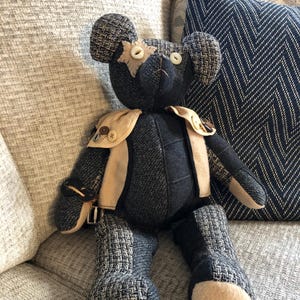 May include: A handcrafted teddy bear with a patchwork design. The bear is mainly dark blue with tweed-like fabric accents and beige felt paws. It has button eyes and a beige felt star on its head. The bear is wearing a beige vest with button details.