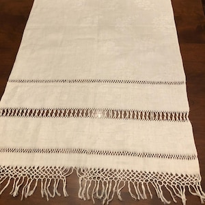 May include: White rectangular table runner with decorative cut-out patterns and fringe trim. The runner features horizontal rows of openwork and a floral pattern in the fabric. A vintage-style home decor item.