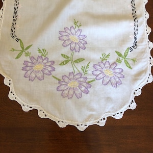 May include: White crocheted table runner with embroidered purple flowers and green leaves. The runner has a scalloped edge.