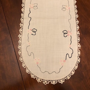 May include: Oval off-white table runner with a crocheted lace edge. The runner features embroidered pink roses and gray and black decorative lines. A delicate, vintage-style home decor item.