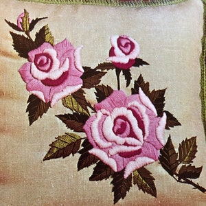 May include: Embroidered pillow with pink roses and green leaves. The roses have a detailed, layered design with a light pink center and darker pink outer petals. The pillow has a light beige background and a green fringe border.