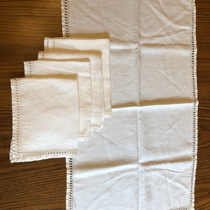 Op de afbeelding: Een set witte linnen servetten en een bijpassend tafelkleed, gerangschikt op een houten oppervlak. De servetten zijn gevouwen en gestapeld, met een decoratieve gehaakte rand. Het tafelkleed is uitgevouwen en toont zijn grootte en een vergelijkbare rand.