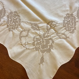 May include: Cream-colored tablecloth with embroidered floral and leaf patterns. The embroidery is in a light beige color, creating a subtle contrast. The edge of the tablecloth has a scalloped design.
