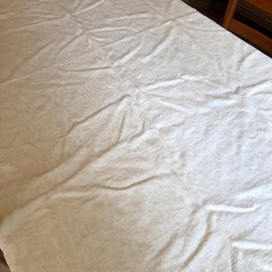 May include: A cream-colored tablecloth draped over a wooden table. The fabric has a subtle textured pattern and visible folds. The table is partially visible, with wooden chairs in the background. This tablecloth is suitable for dining or special events.