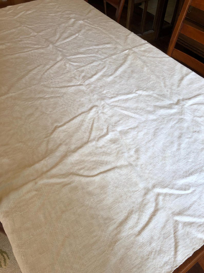 May include: A cream-colored tablecloth draped over a wooden table. The fabric has a subtle textured pattern and visible folds. The table is partially visible, with wooden chairs in the background. This tablecloth is suitable for dining or special events.