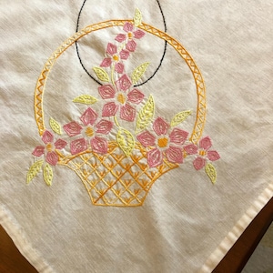 May include: A white linen tablecloth with an embroidered basket of pink, yellow, and orange flowers. The basket is outlined in orange and yellow thread.