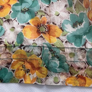 May include: Close-up of a fabric with a floral pattern. The fabric features a white background with orange, yellow, and teal flowers. The fabric has a green seam along the bottom edge.