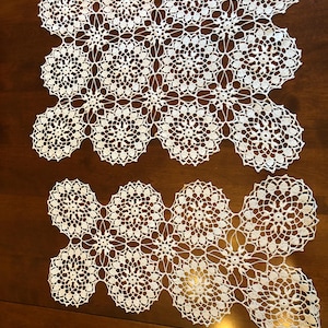 Könnte beinhalten: Zwei weiße, gehäkelte Deckchen mit floralem Muster. Die Deckchen liegen auf einer dunkelbraunen Holzoberfläche. Das filigrane Design weist sich wiederholende kreisförmige Motive und einen gewellten Rand auf, ideal für die Heimdekoration.