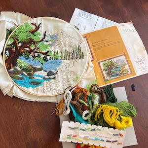 May include: An embroidery kit depicting a landscape with a mountain, trees, and water. The kit includes a completed embroidery piece in a wooden hoop, yarn, and instructions. The Creative Circle brand is visible on the packaging.