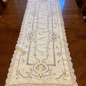 May include: Cream-colored table runner with intricate cutwork and lace edging. The runner features floral and scroll designs, creating an elegant and vintage aesthetic. The runner is placed on a dark wooden table.