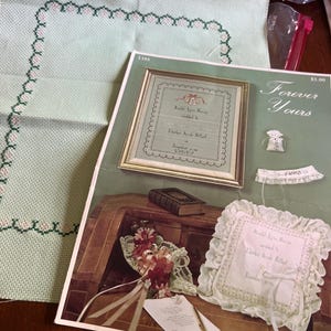 May include: A light green cross-stitch fabric with a pink and green border sits beside a vintage wedding sampler pattern. The pattern is titled "Forever Yours" and features a framed certificate, a ring pillow, and a bouquet.
