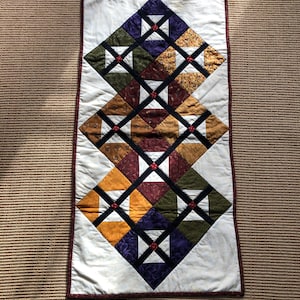 May include: A rectangular quilted table runner with a white background and a geometric pattern of squares in shades of green, purple, brown, and yellow. The squares are outlined in black and have red buttons in the center.