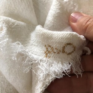 May include: Close-up of a white, textured fabric with frayed edges. The fabric is folded, revealing a glimpse of the word "ROCO" embroidered in gold thread. The material appears soft and delicate.