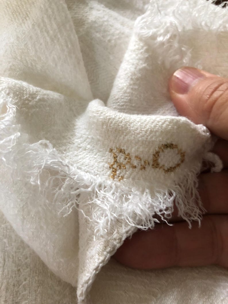 May include: Close-up of a white, textured fabric with frayed edges. The fabric is folded, revealing a glimpse of the word "ROCO" embroidered in gold thread. The material appears soft and delicate.