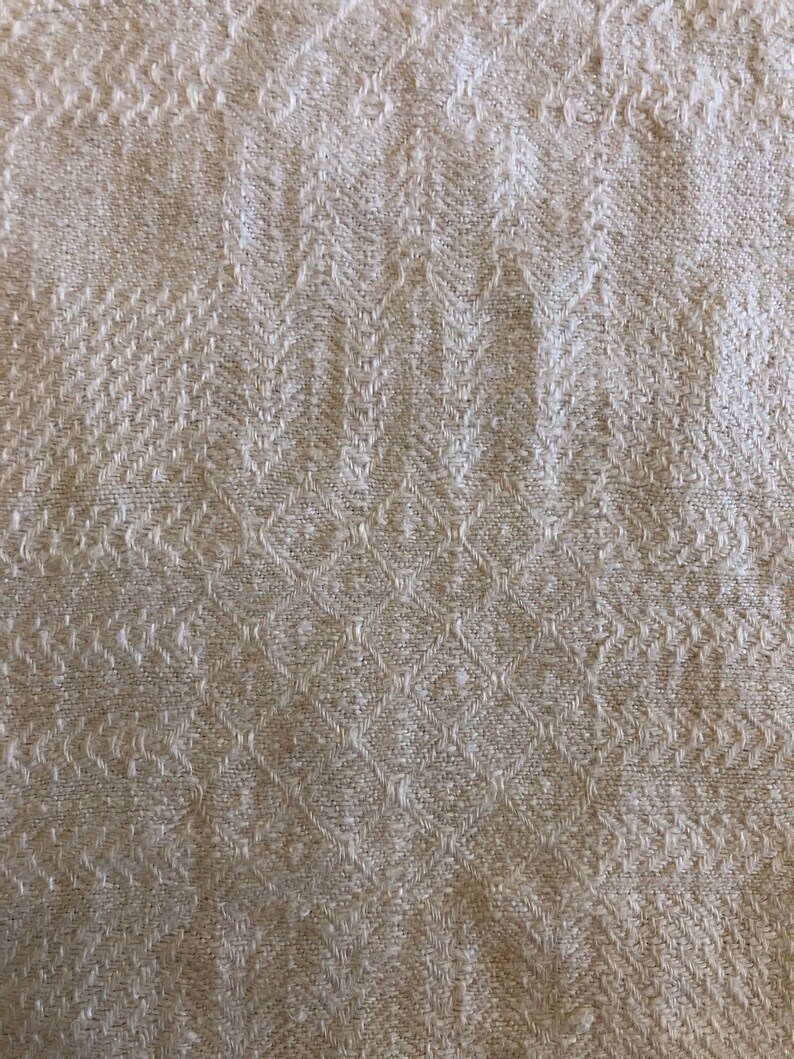 May include: Close-up of a beige textile with a woven pattern. The fabric has a textured surface with a repeating diamond and chevron design. The neutral color and detailed weave suggest a natural fiber material.
