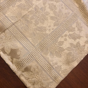 Puede incluir: Un mantel beige con un estampado floral y un borde texturizado. El mantel está hecho de una tela tejida y tiene un brillo sutil. El diseño presenta motivos florales repetidos dentro de una cuadrícula.