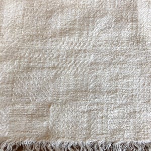 May include: Off-white woven textile with a textured surface and a fringed edge. The fabric has a subtle pattern and a soft, natural appearance. The fringe is made of loose threads, adding a rustic touch to the piece. This textile could be a scarf, shawl, or decorative cloth.