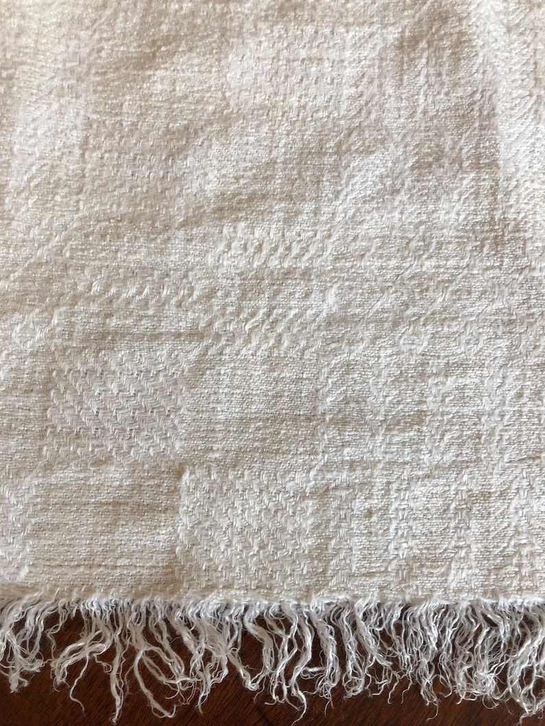 May include: Off-white woven textile with a textured surface and a fringed edge. The fabric has a subtle pattern and a soft, natural appearance. The fringe is made of loose threads, adding a rustic touch to the piece. This textile could be a scarf, shawl, or decorative cloth.