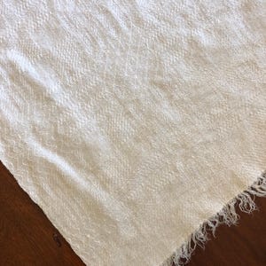 May include: Off-white woven textile with a textured surface and fringed edge. The fabric has a subtle pattern and is draped over a dark wooden surface. The textile appears to be a scarf or shawl.