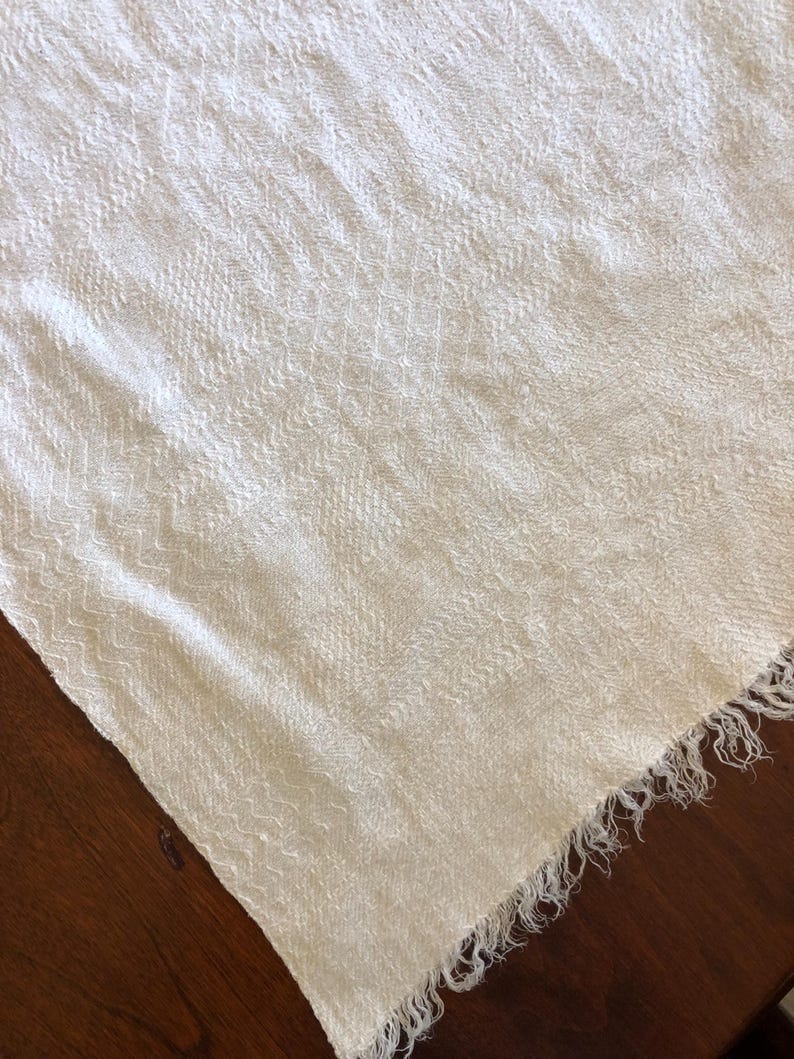 May include: Off-white woven textile with a textured surface and fringed edge. The fabric has a subtle pattern and is draped over a dark wooden surface. The textile appears to be a scarf or shawl.