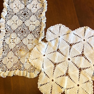 May include: A collection of white, handmade crochet doilies and a rectangular table runner. The table runner has a scalloped edge and a floral pattern. The doilies feature a starburst design. The items are displayed on a dark wooden surface.