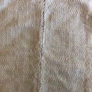 May include: Close-up of a textured, off-white fabric with a repeating pattern. A vertical line of stitching runs down the center. The fabric has a woven appearance, with subtle variations in tone and texture.