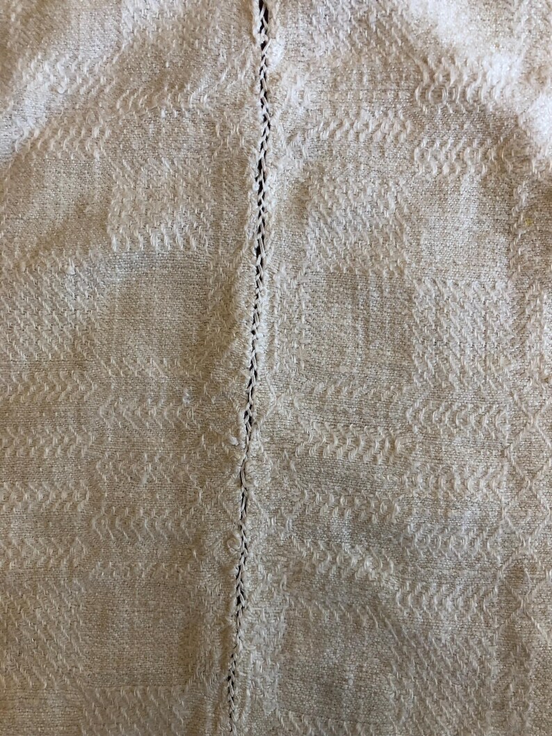 May include: Close-up of a textured, off-white fabric with a repeating pattern. A vertical line of stitching runs down the center. The fabric has a woven appearance, with subtle variations in tone and texture.