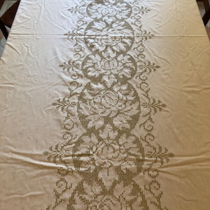 Könnte beinhalten: Cremefarbene Tischdecke mit einem aufwendigen, gestickten Blumenmuster, das in der Mitte verläuft. Die Stickerei ist in einem gedämpften Goldton gehalten und bildet einen Kontrast zum helleren Hintergrund. Der Tisch steht auf einem Holztisch.