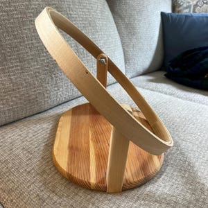 May include: A light-colored wooden embroidery hoop stand featuring a circular frame attached to a vertical support post, which is mounted onto a flat, oval-shaped wooden base with a visible grain pattern and a small metal wing nut fastener.