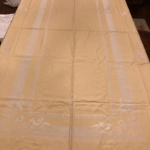 Puede incluir: Un mantel rectangular grande de color amarillo pálido. El mantel presenta un diseño rectangular central con bordes blancos y patrones florales decorativos. La tela parece lisa, posiblemente lino o un material similar.
