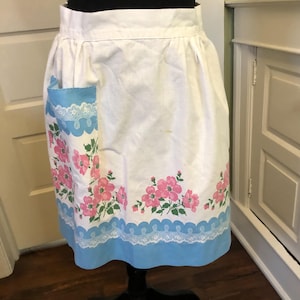 May include: White apron with a blue pocket and a blue scalloped border. The apron features a floral pattern of pink flowers and green leaves. The pocket has a white lace trim.