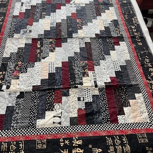 Könnte beinhalten: Eine handgefertigte Steppdecke mit einem Patchwork-Design in Schwarz-, Weiß- und Burgundertönen. Die Steppdecke hat ein geometrisches Muster mit rechteckigen Blöcken und einen Rand mit asiatisch inspiriertem Text. Die Steppdecke liegt auf einem Bett.