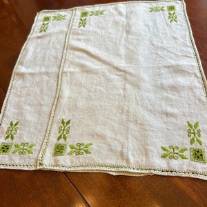 Puede incluir: Dos servilletas de lino blanquecino con diseños florales bordados en verde. Las servilletas tienen un borde de ganchillo verde y detalles bordados en las esquinas. Se colocan sobre una superficie de madera oscura.